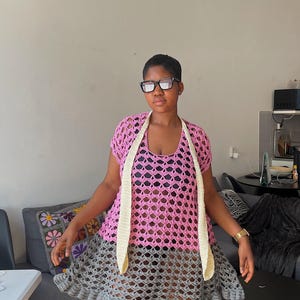 May include: A pink and gray crochet dress with a cream-colored scarf. The dress features a pink top and a gray skirt with a ruffled hem. The open weave design is visible throughout the garment.