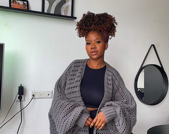 May include: A woman wearing a gray crochet cardigan with a black top and shorts. The cardigan has a loose fit and long sleeves. The woman is standing in front of a white wall with shelves and a mirror.