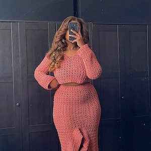 May include: A two-piece crochet outfit in a dusty rose color. The top is a cropped, long-sleeved sweater with a loose fit. The skirt is a long, flowing skirt with a high waist and a ruffled hem.