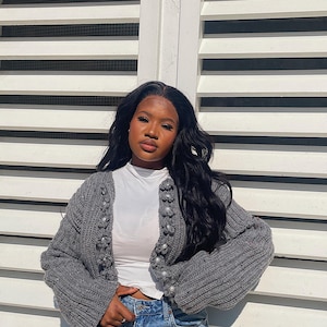 May include: A gray knit cardigan with pearl-like buttons. The cardigan is worn over a white top and paired with denim shorts. The background is a white slatted wall.