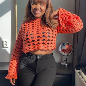 May include: A woman wearing a bright orange crochet top with bell sleeves and black pants.