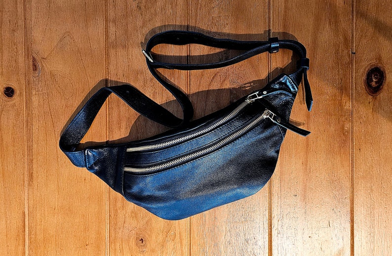 May include: A black leather fanny pack with two zippered compartments and a long adjustable strap.