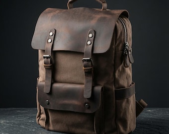 May include: A brown canvas backpack featuring a dark brown leather top flap and a front pocket flap. The bag includes two vertical leather straps with metal buckles, a top handle, and side pockets with leather accents and silver-toned hardware.