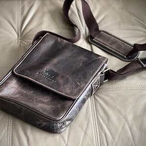 K&ouml;nnte beinhalten: Eine braune Ledertasche mit verstellbarem Schultergurt und einem kleinen Logo mit der Aufschrift "Wildwood" und einer stilisierten Tiersilhouette.