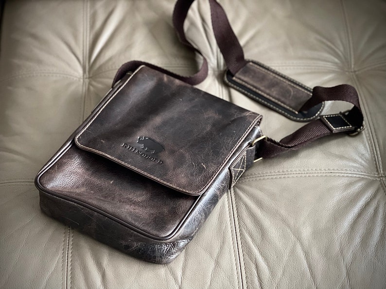 K&ouml;nnte beinhalten: Eine braune Ledertasche mit verstellbarem Schultergurt und einem kleinen Logo mit der Aufschrift "Wildwood" und einer stilisierten Tiersilhouette.