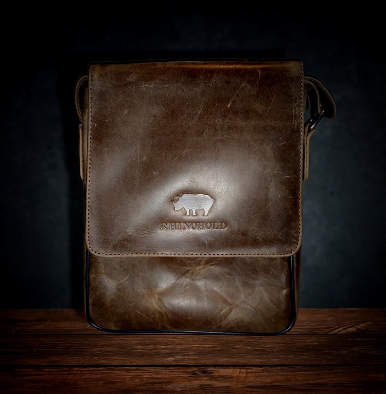 K&ouml;nnte beinhalten: Eine braune Ledertasche mit einem Nashorn-Logo und dem Schriftzug "RHINO HOLD" auf der Vorderseite.