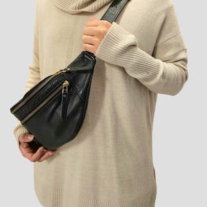 May include: A black leather fanny pack with a gold zipper and strap. The fanny pack is worn over a beige sweater.