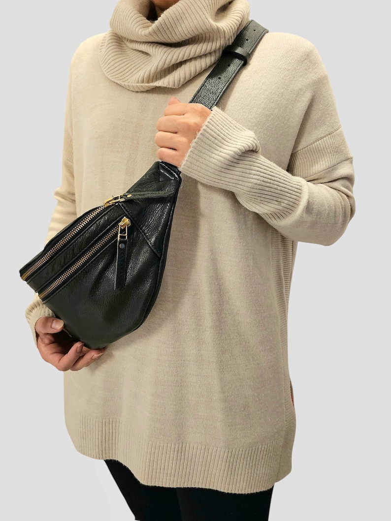 May include: A black leather fanny pack with a gold zipper and strap. The fanny pack is worn over a beige sweater.