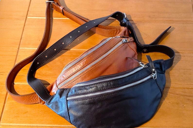 May include: A brown leather fanny pack with a black leather fanny pack attached to it. Both have silver zippers.