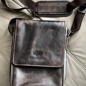 K&ouml;nnte beinhalten: Eine braune Ledertasche mit einem Klappverschluss und einem verstellbaren Schultergurt. Die Tasche hat ein Nashorn-Logo mit dem Text "RhinoHold" darunter.