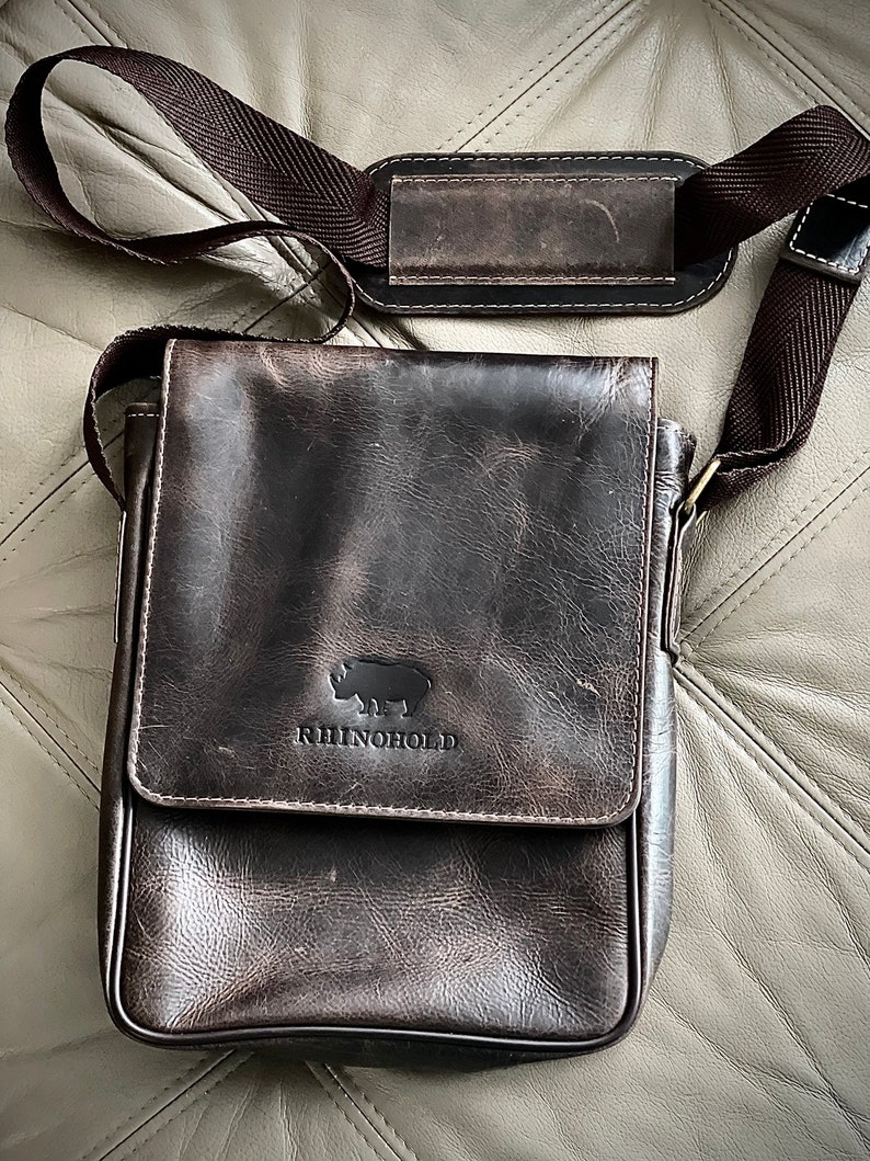 K&ouml;nnte beinhalten: Eine braune Ledertasche mit einem Klappverschluss und einem verstellbaren Schultergurt. Die Tasche hat ein Nashorn-Logo mit dem Text "RhinoHold" darunter.