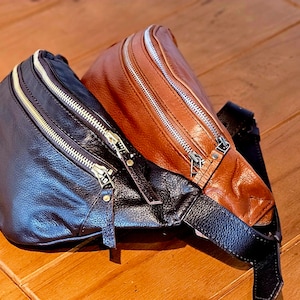 May include: Two leather fanny packs, one brown and one black, with gold zippers. The brown fanny pack is on top of the black fanny pack.