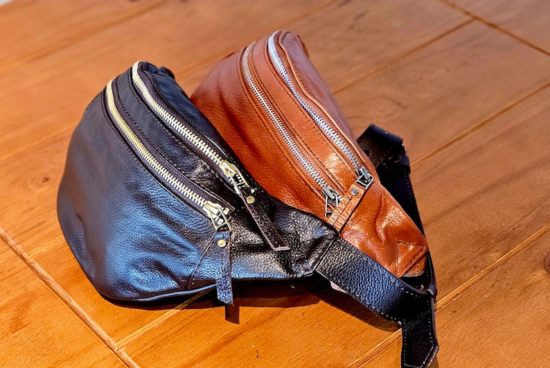 May include: Two leather fanny packs, one brown and one black, with gold zippers. The brown fanny pack is on top of the black fanny pack.
