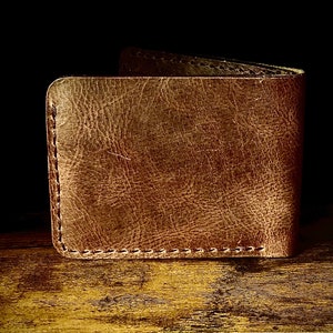 Peut inclure: Un portefeuille en cuir marron avec une finition vieillie et des coutures visibles. Le portefeuille est ferm&eacute; et pos&eacute; sur une surface en bois.