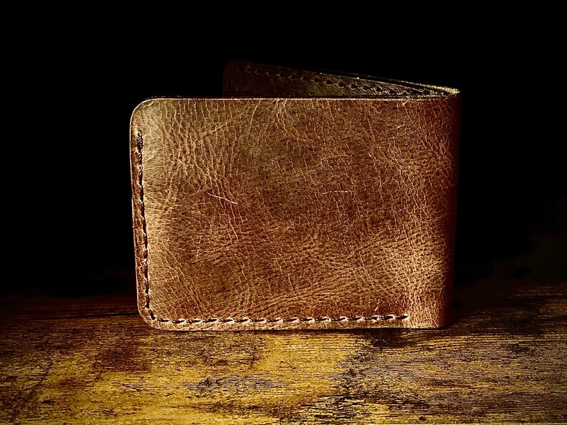 Peut inclure: Un portefeuille en cuir marron avec une finition vieillie et des coutures visibles. Le portefeuille est ferm&eacute; et pos&eacute; sur une surface en bois.