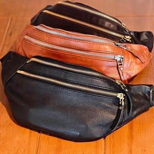 May include: Three leather fanny packs in black, brown, and tan with gold zippers. The fanny packs are stacked on top of each other.