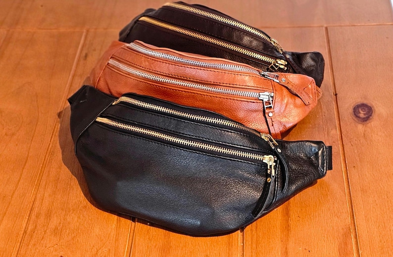 May include: Three leather fanny packs in black, brown, and tan with gold zippers. The fanny packs are stacked on top of each other.