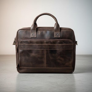 May include: A dark brown leather briefcase featuring a rectangular shape, two top handles, and a front zippered pocket. The bag has visible stitching details, a main zipper closure, and metal hardware attachments on the sides for a shoulder strap.