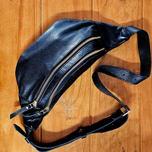 May include: A black leather fanny pack with two gold zippers and a black adjustable strap.