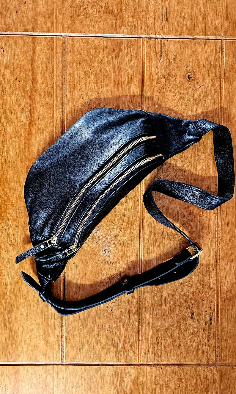 May include: A black leather fanny pack with two gold zippers and a black adjustable strap.