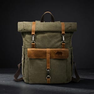 May include: An olive green canvas roll-top backpack featuring tan leather straps, silver-toned metal hardware, and a front pocket flap embossed with a rhinoceros silhouette and the text RHINOGOLD. The bag includes a top handle and side pockets.