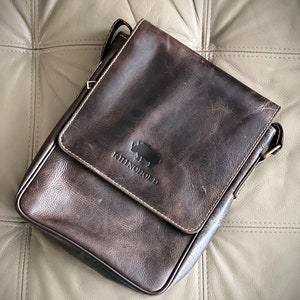 K&ouml;nnte beinhalten: Eine dunkelbraune Ledertasche mit einem Klappverschluss und einem Nashorn-Logo mit der Aufschrift "RhinoHold".