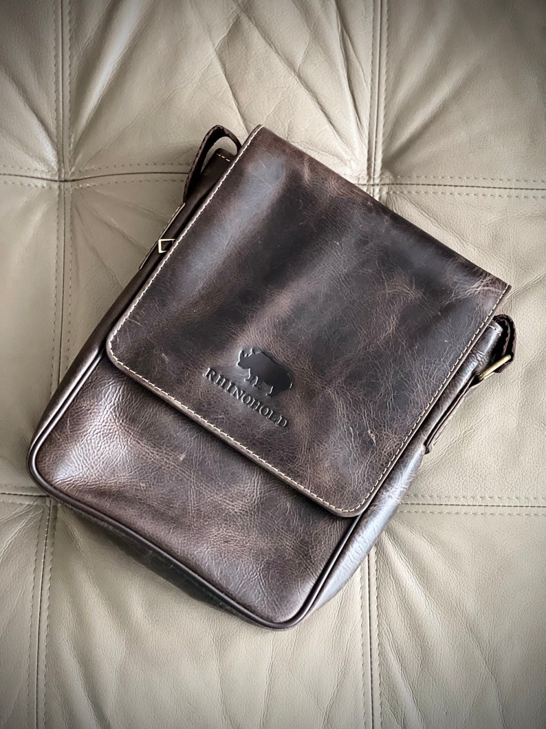 K&ouml;nnte beinhalten: Eine dunkelbraune Ledertasche mit einem Klappverschluss und einem Nashorn-Logo mit der Aufschrift "RhinoHold".