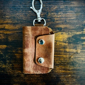 May include: A brown leather key holder with a silver-colored clasp and two silver snap closures. The key holder is made of distressed leather and is designed to hold keys securely. The key holder is shown against a dark wood grain background.