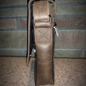 K&ouml;nnte beinhalten: Eine braune Ledertasche mit einem Klappverschluss und einer Rei&szlig;verschlusstasche. Die Tasche hat einen Trageriemen und eine silberne Schnalle.