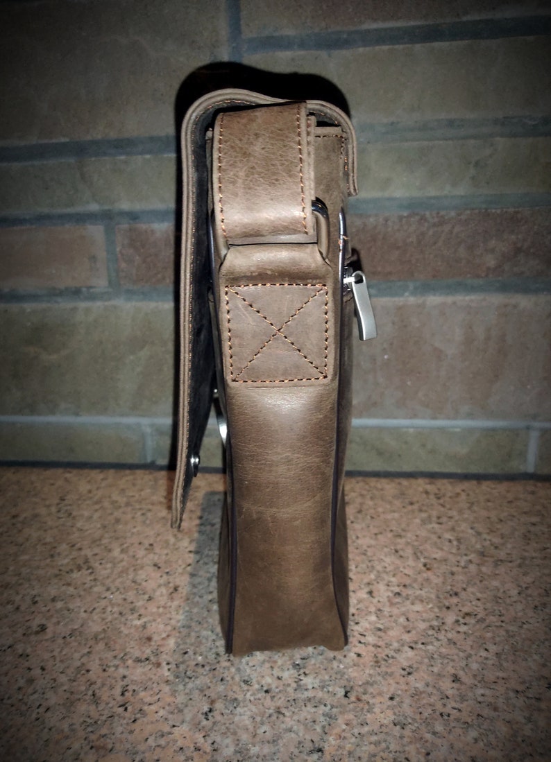 K&ouml;nnte beinhalten: Eine braune Ledertasche mit einem Klappverschluss und einer Rei&szlig;verschlusstasche. Die Tasche hat einen Trageriemen und eine silberne Schnalle.