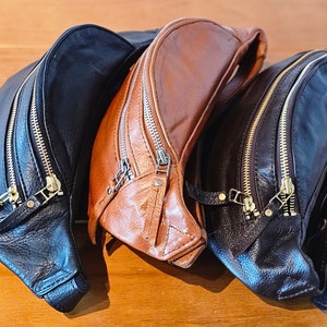 May include: Three leather fanny packs in black and brown. The brown fanny pack has a zipper closure and a flap with a buckle. The black fanny packs have two zipper closures.