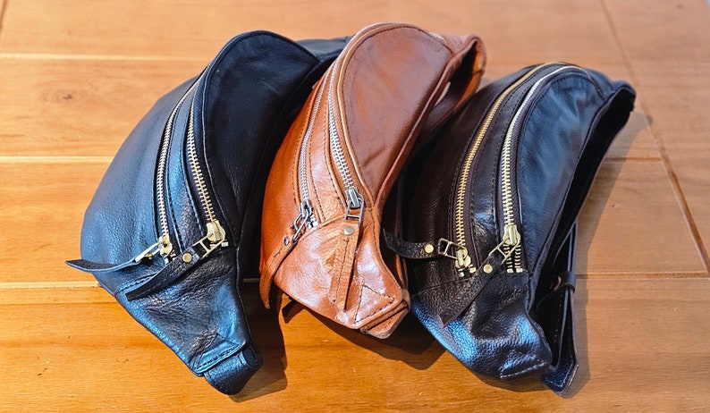 May include: Three leather fanny packs in black and brown. The brown fanny pack has a zipper closure and a flap with a buckle. The black fanny packs have two zipper closures.