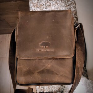 K&ouml;nnte beinhalten: Eine braune Ledertasche mit einem Nashorn-Logo und dem Text "RhinoHold" auf der Vorderseite.