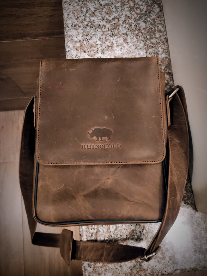 K&ouml;nnte beinhalten: Eine braune Ledertasche mit einem Nashorn-Logo und dem Text "RhinoHold" auf der Vorderseite.