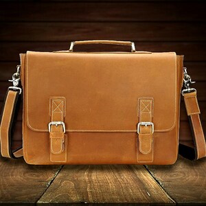 May include: A brown leather briefcase with a top handle and a shoulder strap. The briefcase has two buckles on the front and a flap that closes with a buckle.
