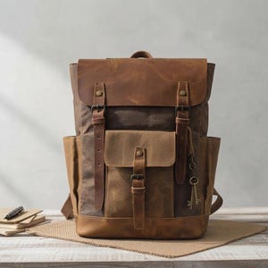 Peut inclure: Un sac à dos en cuir et toile marron doté d'un rabat supérieur avec deux sangles verticales en cuir, une poche frontale centrale avec fermeture à boucle, des poches latérales et une breloque clé en métal suspendue à une boucle.