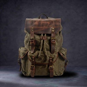 May include: An olive green canvas backpack featuring a dark brown leather top flap and trim. The bag includes two front pockets with leather straps, brass-toned metal buckles, and a drawstring closure under the main flap with a top handle.