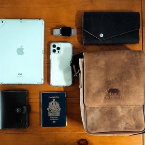 K&ouml;nnte beinhalten: Eine braune Leder-Messenger-Tasche mit einem Nashorn-Logo, eine schwarze Ledergeldb&ouml;rse, ein wei&szlig;es iPad, ein wei&szlig;es iPhone, ein kanadischer Reisepass, eine schwarze Ledergeldb&ouml;rse und eine silberne Smartwatch.