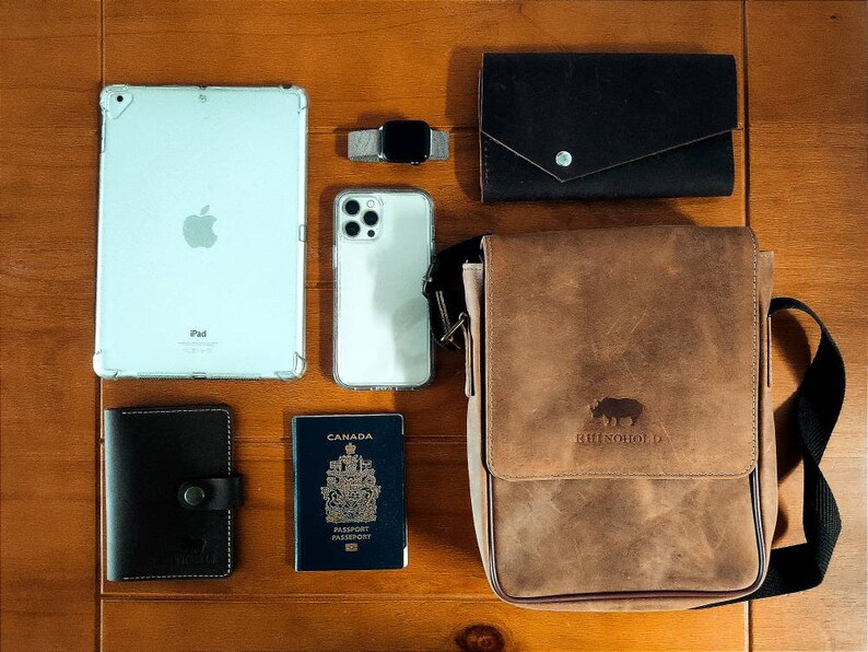 K&ouml;nnte beinhalten: Eine braune Leder-Messenger-Tasche mit einem Nashorn-Logo, eine schwarze Ledergeldb&ouml;rse, ein wei&szlig;es iPad, ein wei&szlig;es iPhone, ein kanadischer Reisepass, eine schwarze Ledergeldb&ouml;rse und eine silberne Smartwatch.