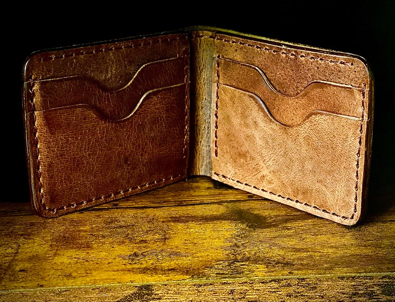 Peut inclure: Un portefeuille en cuir marron avec deux fentes pour cartes de chaque c&ocirc;t&eacute;, ouvert et pos&eacute; sur une surface en bois.