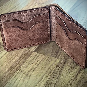 Peut inclure: Un portefeuille en cuir marron avec plusieurs emplacements pour cartes et un porte-billets. Le portefeuille est ouvert et l'int&eacute;rieur est visible.