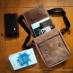 K&ouml;nnte beinhalten: Eine braune Leder-Messenger-Tasche mit Klappenverschluss und einem langen, verstellbaren Trageriemen. Die Tasche ist offen und enth&auml;lt einen Reisepass, ein Telefon und ein Buch mit dem Titel "GRIT: The Power of Passion and Perseverance" von Angela Duckworth.