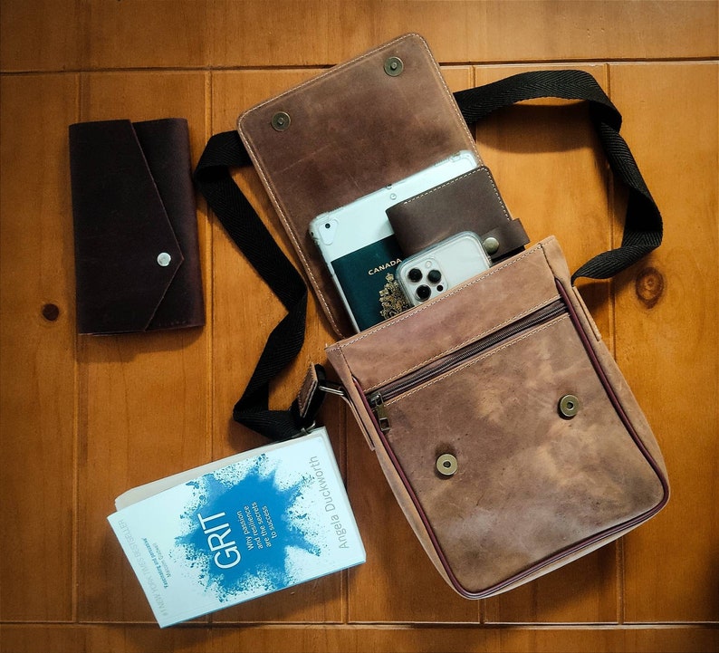 K&ouml;nnte beinhalten: Eine braune Leder-Messenger-Tasche mit Klappenverschluss und einem langen, verstellbaren Trageriemen. Die Tasche ist offen und enth&auml;lt einen Reisepass, ein Telefon und ein Buch mit dem Titel "GRIT: The Power of Passion and Perseverance" von Angela Duckworth.