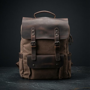 May include: A brown canvas backpack featuring dark brown leather accents, a top handle, and a fold-over flap secured with two adjustable buckle straps. The front includes a horizontal leather pocket with metal snap buttons and side pockets.