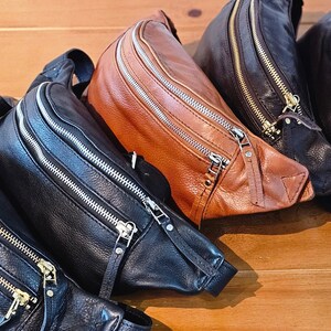 May include: Four leather fanny packs in black and brown with gold zippers. The fanny packs are arranged in a row, with the brown one in the front.
