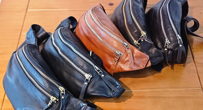 May include: Four leather fanny packs in black and brown with gold zippers. The fanny packs are arranged in a row, with the brown one in the front.