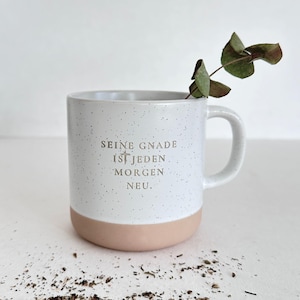 Könnte beinhalten: Eine weiße Keramiktasse mit gesprenkeltem Design und hellrosa Boden. Die Tasse hat einen Henkel und trägt den Text "SEINE GNADE IST JEDEN MORGEN NEU." Ein Zweig Grünzeug befindet sich in der Tasse.