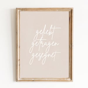Könnte beinhalten: Ein gerahmter Druck mit hellbraunem Hintergrund und weißem Text, der "geliebt getragen gesegnet" lautet.