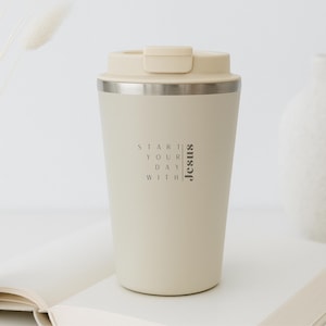 Könnte beinhalten: Ein cremefarbener Reisebecher mit passendem Deckel und silbernem Band. Auf dem Becher steht der Text "Start Your Day With Jesus". Der Becher steht auf einer weißen Oberfläche.