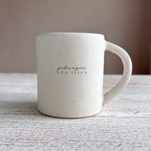 Könnte beinhalten: Weiße Keramiktasse mit gesprenkeltem Finish und schwarzem Text, der "getragen von Jesus" lautet.